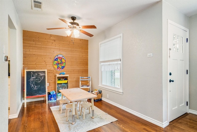 recreation room featuring a ceiling fan, visible vents, wood walls, wood finished floors, and baseboards