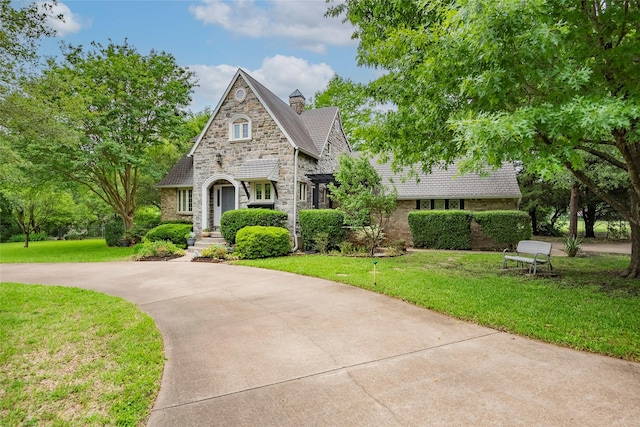 tudor house featuring a chimney, brick siding, curved driveway, a front lawn, and stone siding