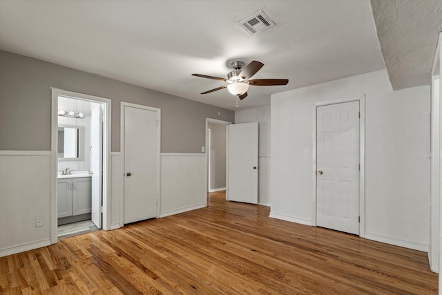 unfurnished bedroom featuring connected bathroom, ceiling fan, wainscoting, light wood finished floors, and visible vents