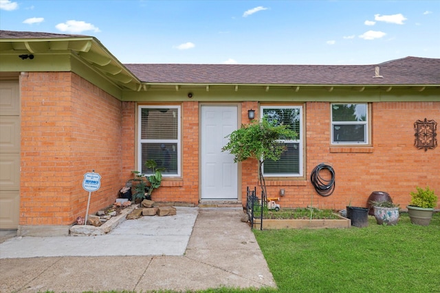 doorway to property featuring a shingled roof, a yard, and brick siding