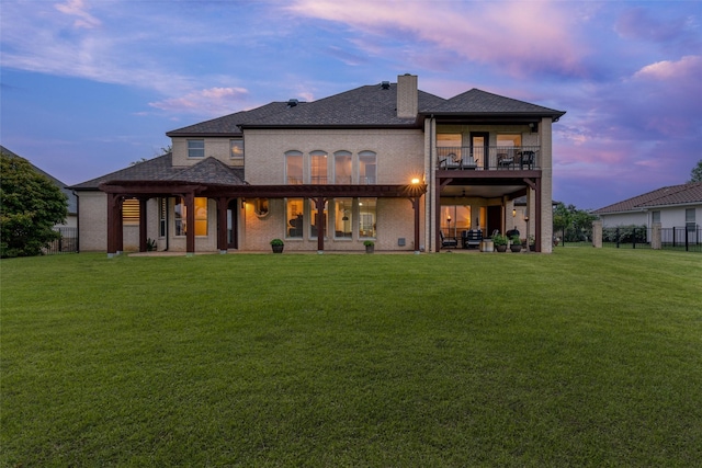 back of property at dusk with a chimney, brick siding, fence, a balcony, and a yard