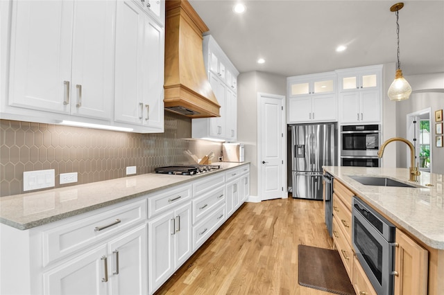 kitchen featuring glass insert cabinets, custom range hood, a sink, light wood-style flooring, and stainless steel appliances