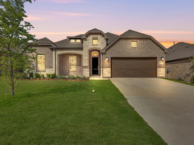 french country inspired facade featuring a shingled roof, an attached garage, brick siding, a lawn, and driveway