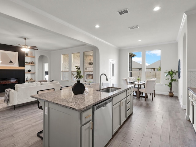 kitchen featuring arched walkways, stainless steel dishwasher, a sink, light wood finished floors, and an island with sink