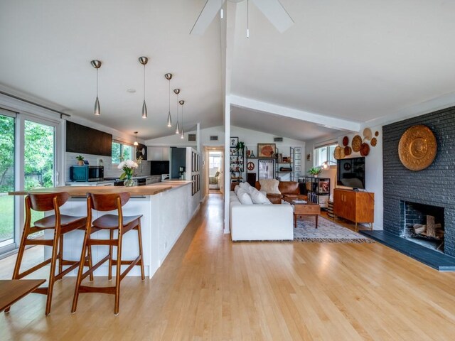living area featuring ceiling fan, lofted ceiling with beams, a brick fireplace, and light wood-type flooring