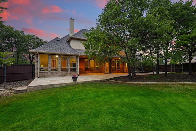 back of house at dusk featuring fence, a lawn, a patio area, and roof with shingles