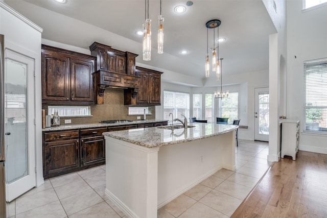 kitchen with a kitchen island with sink, dark brown cabinetry, light stone countertops, a sink, and a chandelier