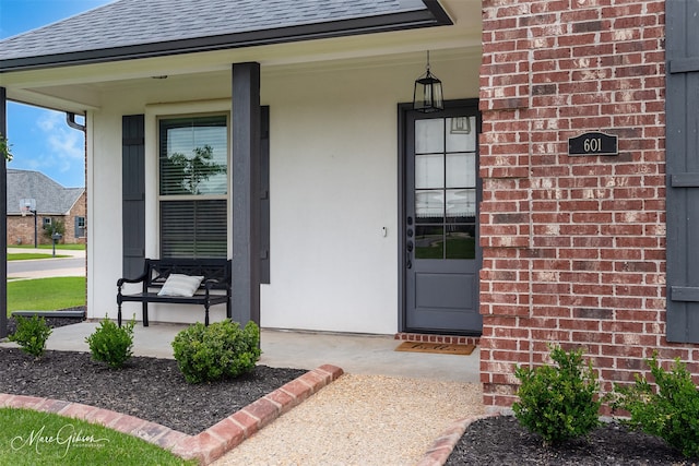 view of exterior entry with a shingled roof, a porch, and brick siding
