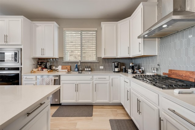 kitchen with light wood-style floors, wall chimney exhaust hood, white cabinetry, a sink, and appliances with stainless steel finishes