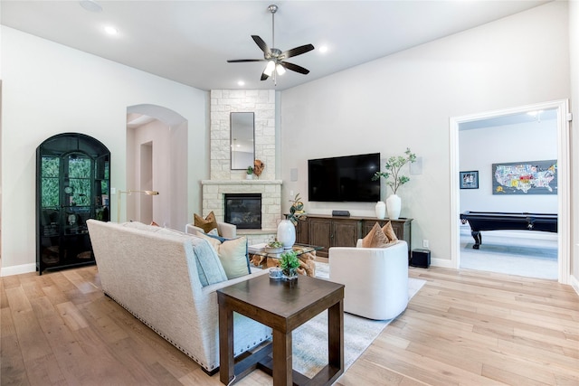 living room with ceiling fan, a stone fireplace, arched walkways, baseboards, and light wood-style floors