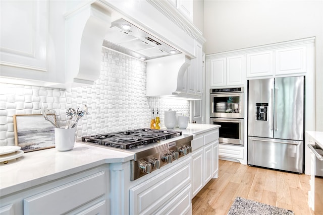 kitchen with backsplash, appliances with stainless steel finishes, white cabinets, custom exhaust hood, and light wood finished floors
