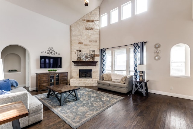 living room featuring a stone fireplace, baseboards, dark wood-style floors, high vaulted ceiling, and arched walkways