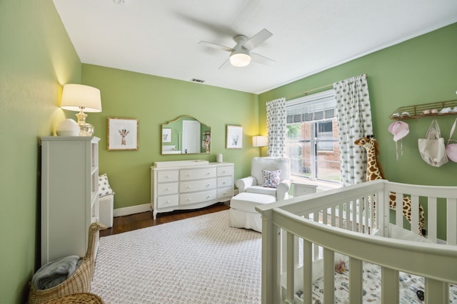 bedroom with visible vents, wood finished floors, baseboards, a crib, and a ceiling fan