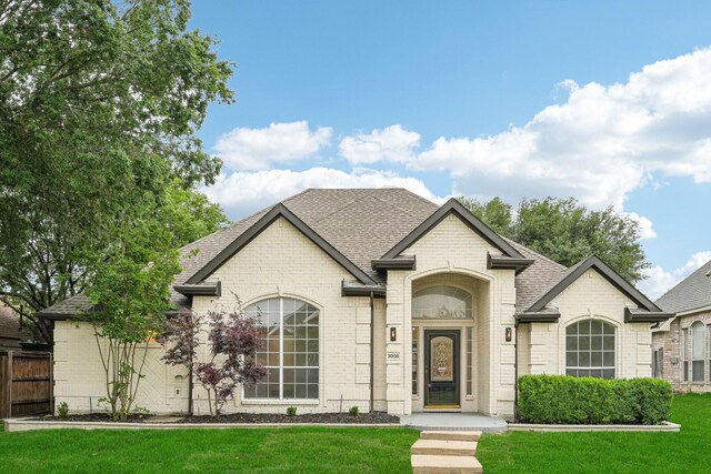 french provincial home with roof with shingles, fence, a front yard, and brick siding