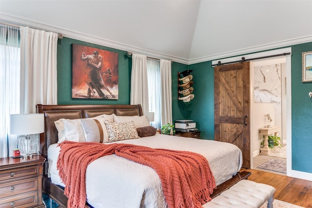 bedroom featuring wood finished floors, baseboards, a barn door, ensuite bath, and vaulted ceiling