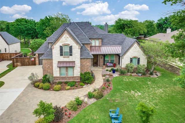 french country inspired facade featuring a standing seam roof, a chimney, a front yard, fence, and stone siding