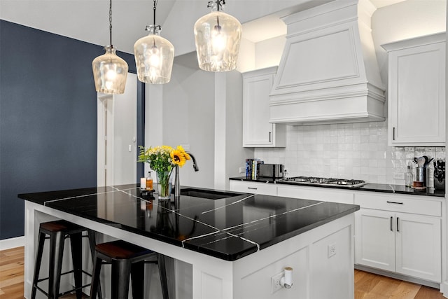 kitchen featuring a kitchen island with sink, dark countertops, tasteful backsplash, stainless steel gas cooktop, and custom exhaust hood