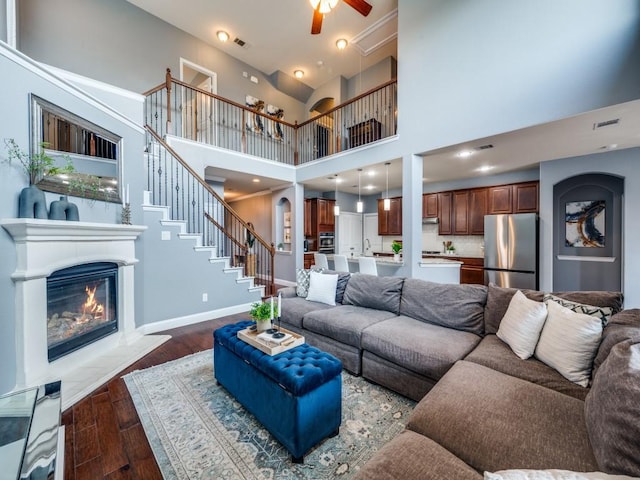 living room with a glass covered fireplace, dark wood finished floors, a ceiling fan, visible vents, and stairway