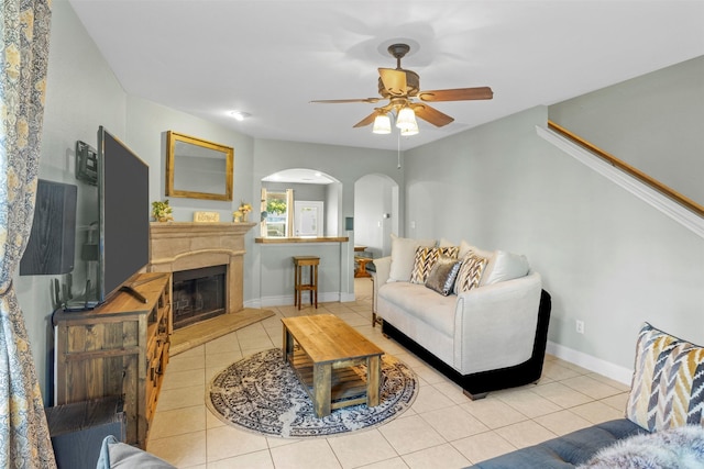 living room with ceiling fan, arched walkways, a glass covered fireplace, baseboards, and light tile patterned floors