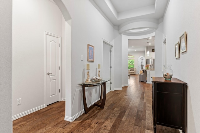 hall featuring dark wood-style flooring, arched walkways, baseboards, and a tray ceiling