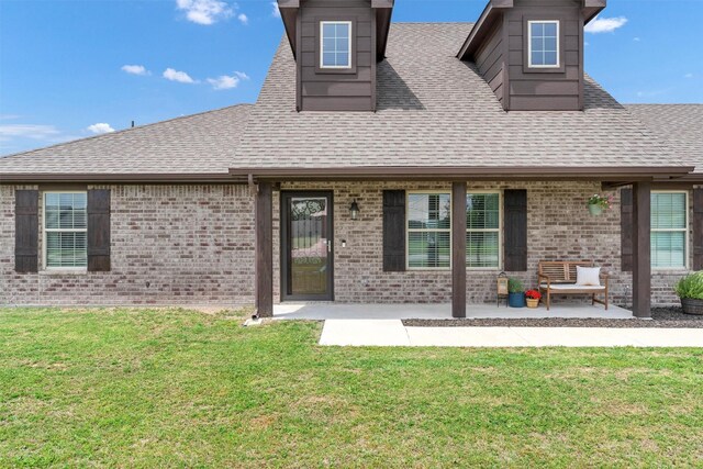 rear view of property featuring a yard, brick siding, a patio, and roof with shingles