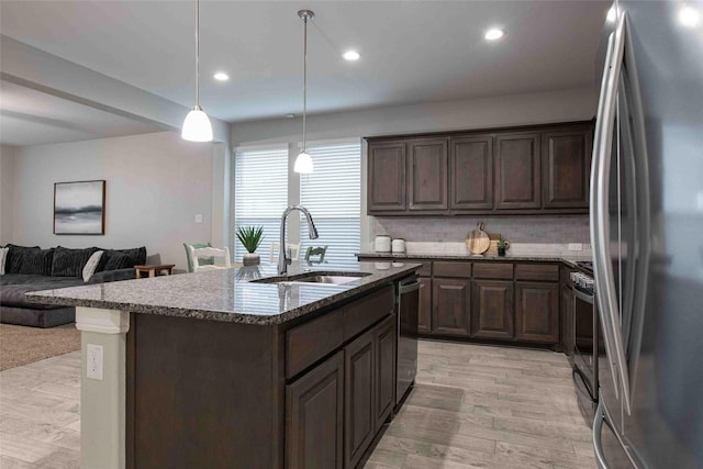 kitchen with dark brown cabinetry, a sink, dark stone counters, stainless steel appliances, and open floor plan