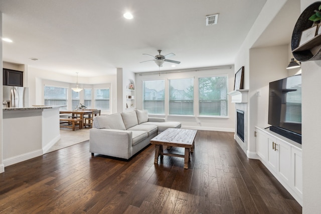 living area featuring a ceiling fan, visible vents, baseboards, a glass covered fireplace, and dark wood-type flooring