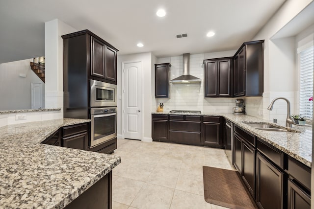 kitchen featuring wall chimney range hood, light stone countertops, a sink, stainless steel appliances, and backsplash