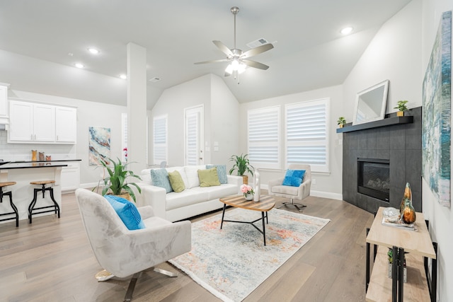 living room with a ceiling fan, visible vents, a tiled fireplace, vaulted ceiling, and light wood-type flooring