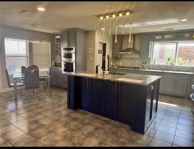 kitchen featuring decorative backsplash, light stone countertops, an island with sink, wall chimney range hood, and stainless steel appliances