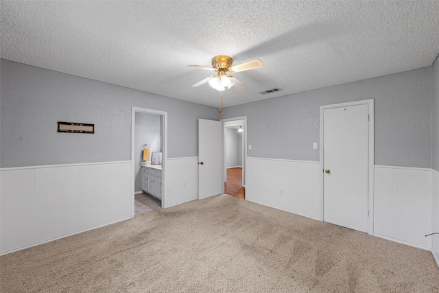 unfurnished bedroom featuring connected bathroom, light colored carpet, a textured ceiling, a wainscoted wall, and visible vents