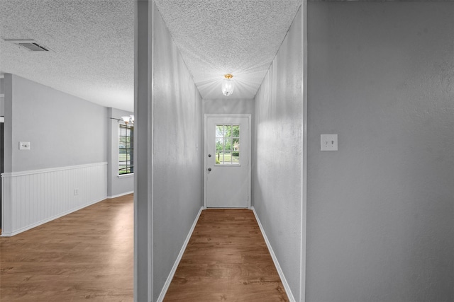 hallway with a textured ceiling, visible vents, wood finished floors, baseboards, and a chandelier