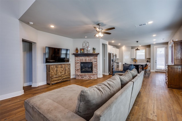 living room with ceiling fan, a brick fireplace, baseboards, wood finished floors, and visible vents
