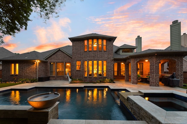 back of property at dusk featuring brick siding, a patio area, a pool with connected hot tub, and a chimney