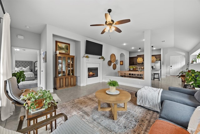 living area featuring ceiling fan, arched walkways, baseboards, light tile patterned floors, and a lit fireplace