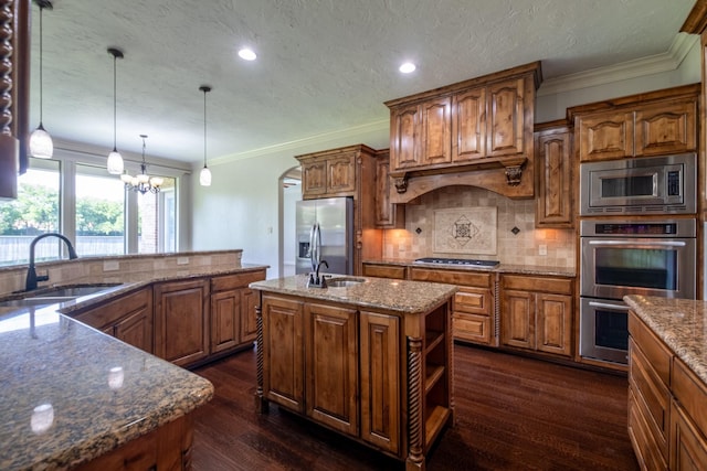 kitchen featuring brown cabinetry, an island with sink, tasteful backsplash, a sink, and appliances with stainless steel finishes