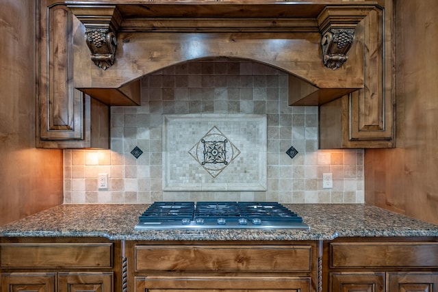 kitchen with stainless steel gas cooktop, backsplash, brown cabinets, and dark stone countertops