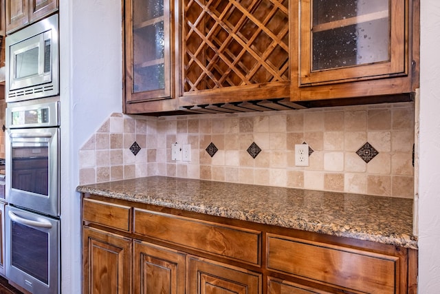 kitchen featuring brown cabinetry, dark stone countertops, stainless steel appliances, tasteful backsplash, and glass insert cabinets