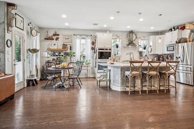 kitchen with dark wood-style floors, a breakfast bar, stainless steel appliances, wall chimney exhaust hood, and an island with sink