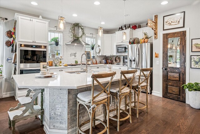 kitchen featuring wall chimney range hood, a kitchen island with sink, stainless steel appliances, decorative backsplash, and dark wood-style floors