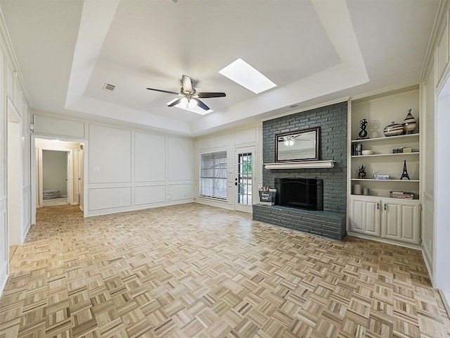 unfurnished living room with a ceiling fan, visible vents, a skylight, a brick fireplace, and a tray ceiling