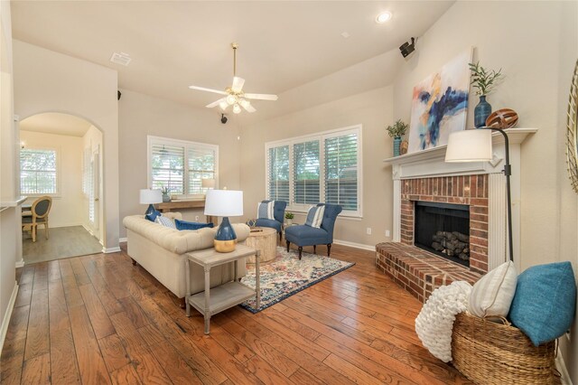 living area with wood-type flooring, a ceiling fan, a brick fireplace, baseboards, and arched walkways