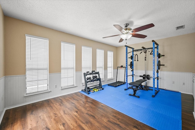 exercise room featuring a textured ceiling, ceiling fan, wainscoting, and wood finished floors