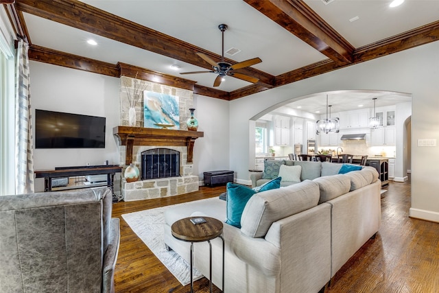 living room with dark wood-style flooring, beamed ceiling, a stone fireplace, arched walkways, and ceiling fan with notable chandelier