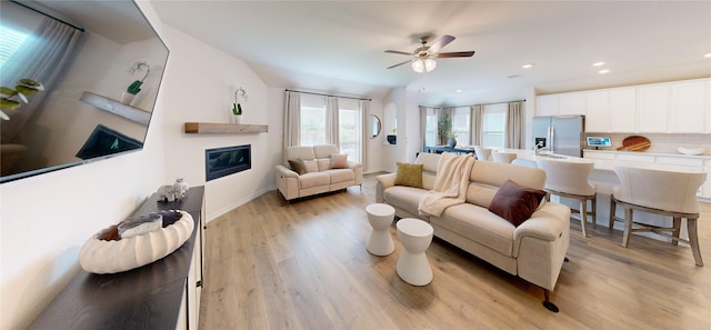 living area with a glass covered fireplace, light wood-style floors, baseboards, ceiling fan, and recessed lighting