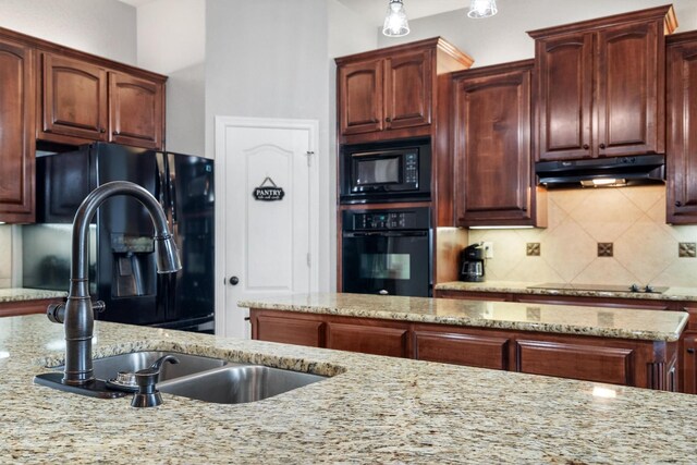 kitchen featuring light stone countertops, under cabinet range hood, a sink, backsplash, and black appliances