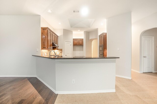 kitchen with a peninsula, dark countertops, under cabinet range hood, brown cabinets, and arched walkways