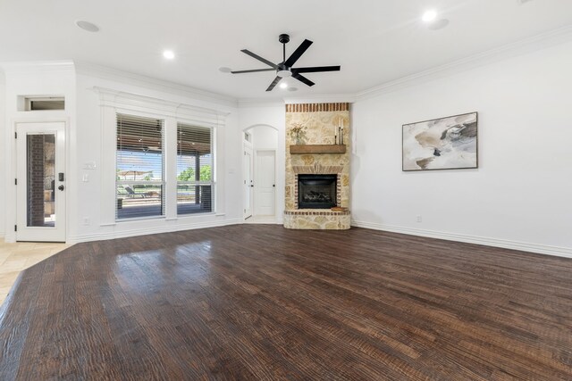 unfurnished living room with a ceiling fan, a stone fireplace, wood finished floors, baseboards, and ornamental molding