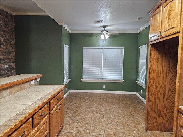 kitchen featuring a ceiling fan, visible vents, baseboards, ornamental molding, and tile counters