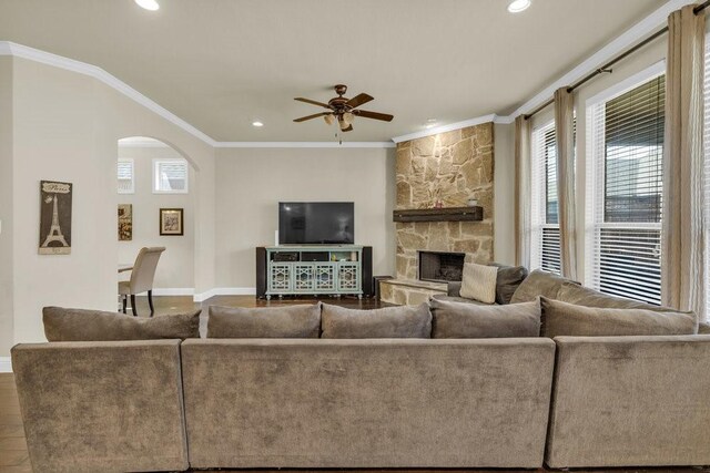 living area with arched walkways, a stone fireplace, a ceiling fan, baseboards, and ornamental molding